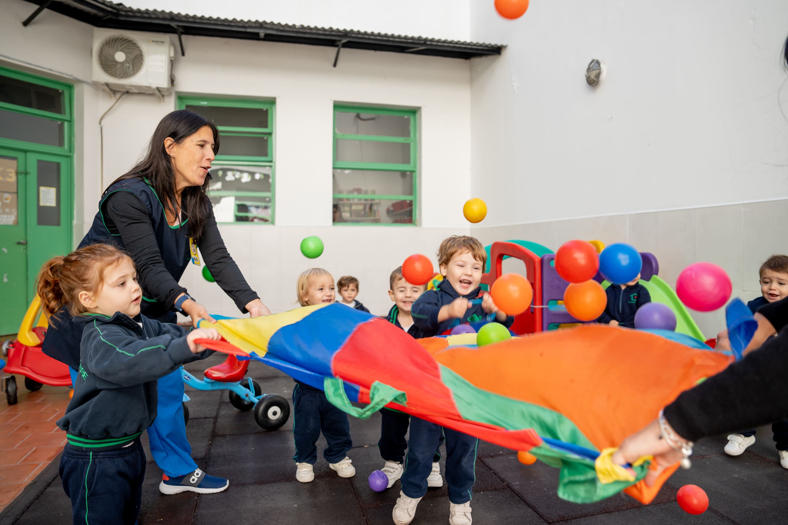 Maestra y nenes jugando con un paracaídas de colores lleno de pelotas en el patio de The Green House Kindergarten