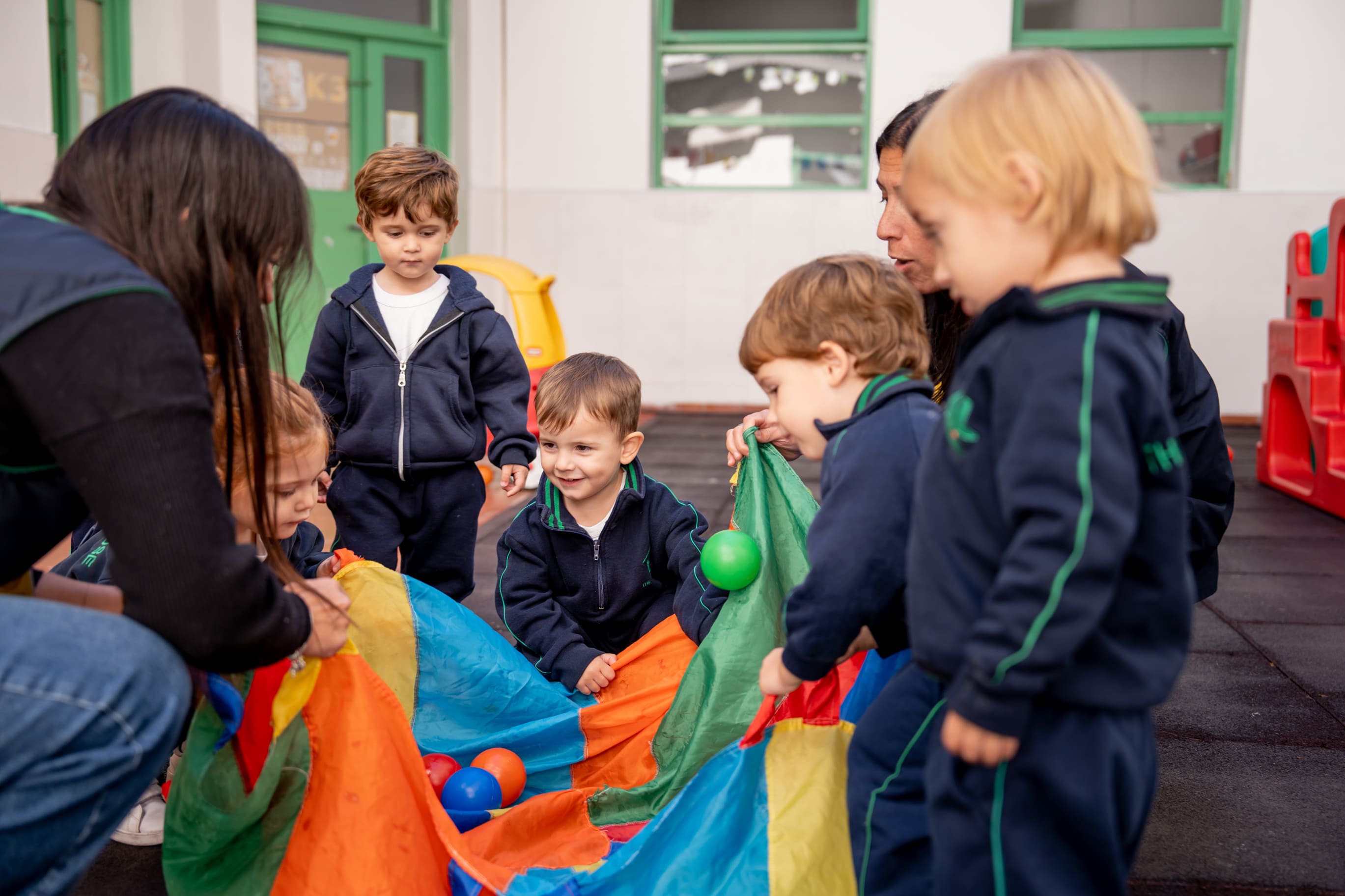 Maestras y nenes jugando con un paracaídas de colores y pelotas en el patio de The Green House Kindergarten